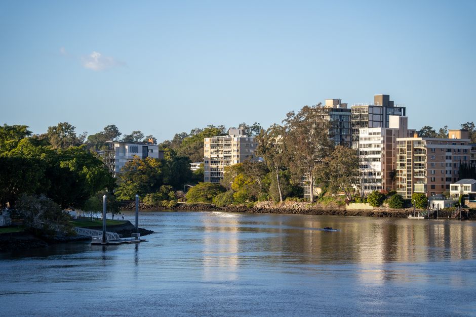A scenic view of multiple modern residential buildings situated along the riverbank in Richmond, with lush green trees in the foreground and background. The river reflects the clear blue sky above, an